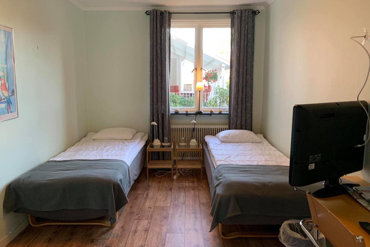 Een kamer met twee eenpersoonsbedden, een raam met gordijnen, een radiator, een bureau met een computer en een prullenbak.