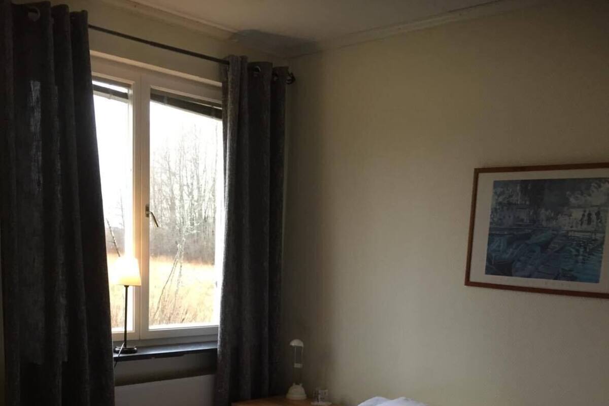 Hotelkamer met een bed, nachtkastje, lamp, raam met gordijnen en een ingelijst schilderij aan de muur.