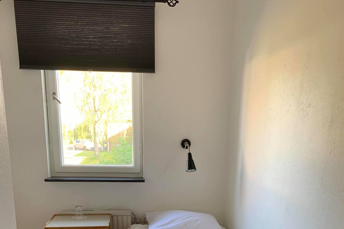 Een kleine, eenvoudig ingerichte kamer met een enkel bed, een tafeltje, een radiator en een raam met uitzicht op groen.