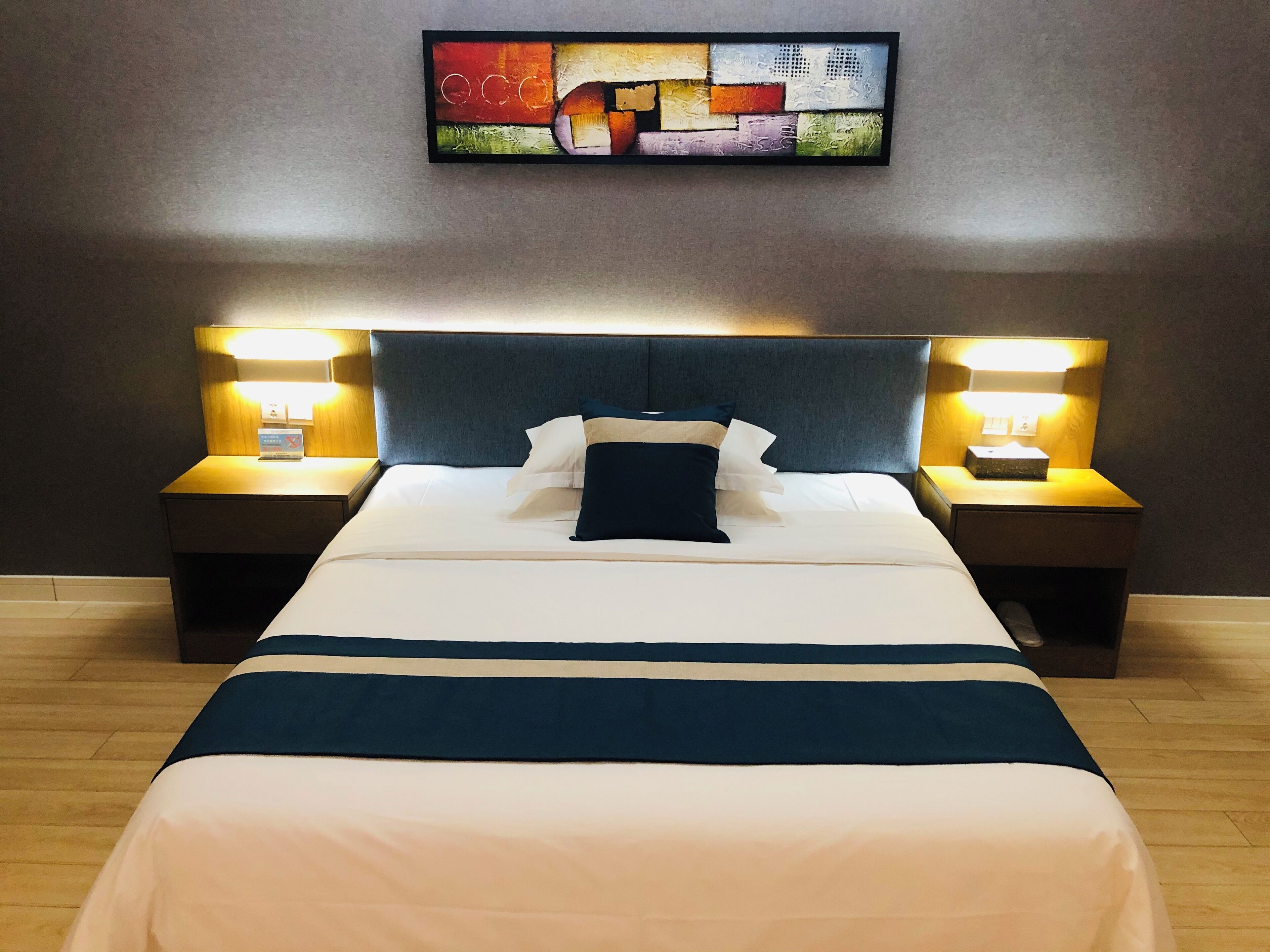 A neatly made bed with a dark headboard, two bedside tables with lamps, and a colorful abstract painting on the wall.