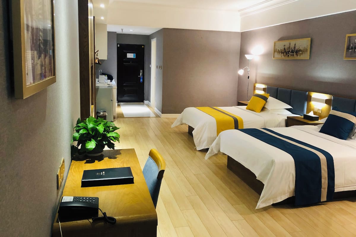 Habitación de hotel con dos camas, un escritorio con una computadora, una silla, una planta y una puerta que da a un pasillo.