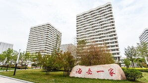 Un complejo moderno de edificios con varias estructuras altas, un área ajardinada con árboles y una piedra con caracteres chinos.