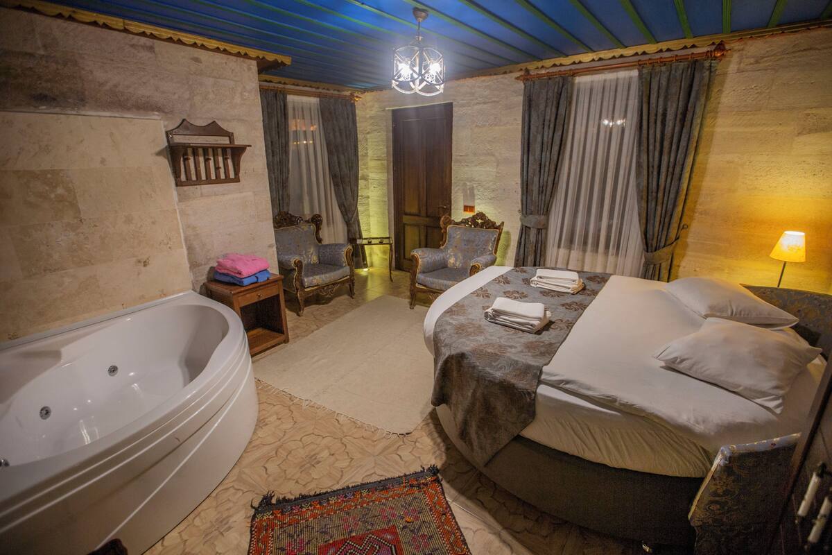 A room with a large bathtub, a bed with pillows, a chair, and a small table with a lamp.