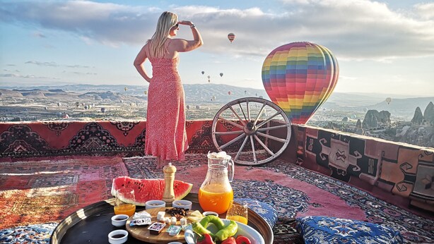 A terrace with a view of hot air balloons, a woman in a red dress, and a table with food and drinks. - Hermes Cave Hotel (Nevsehir)