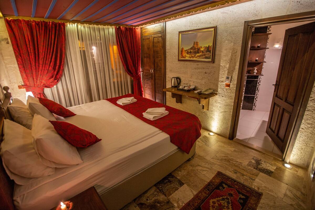 A hotel room with a large bed, red curtains, a painting on the wall, a small shelf with a kettle, and a wooden door.