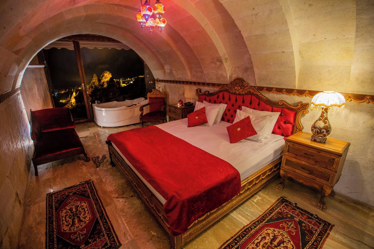 A room with a bed, a chair, a nightstand, and a view of a cityscape through an arched doorway.