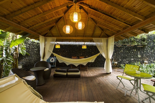 A covered outdoor seating area with wicker furniture, a hammock, and a stone wall backdrop.