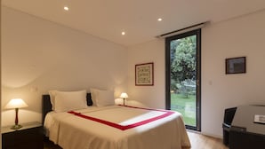 Un dormitorio con una cama grande, dos lámparas de noche, un escritorio y un ventanal corredero hacia un jardín.