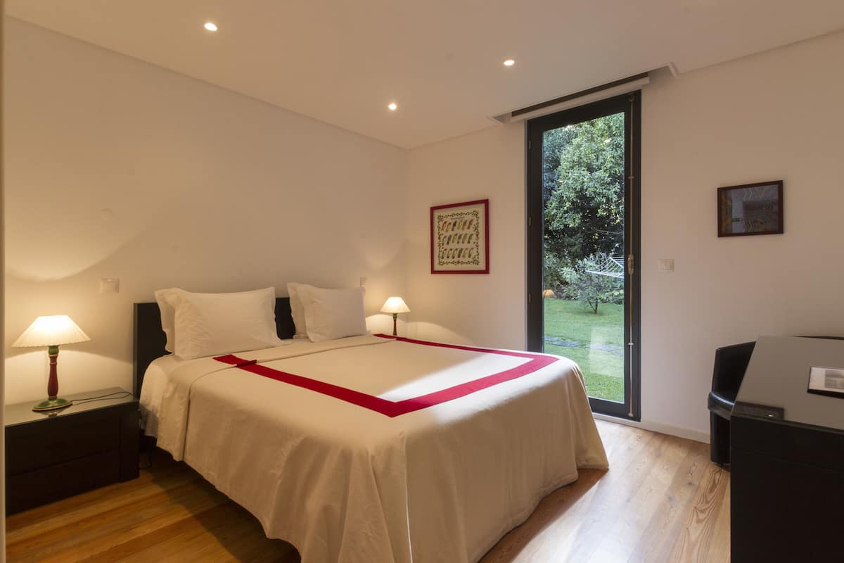 A bedroom with a large bed, two bedside lamps, a desk, and a sliding glass door to a garden.
