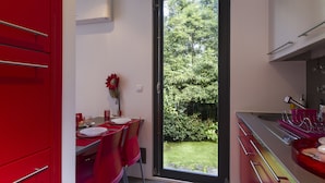 Una cocina moderna con armarios rojos, un comedor con una mesa dispuesta para dos personas, y vistas a un jardín exuberante a través de un ventanal.