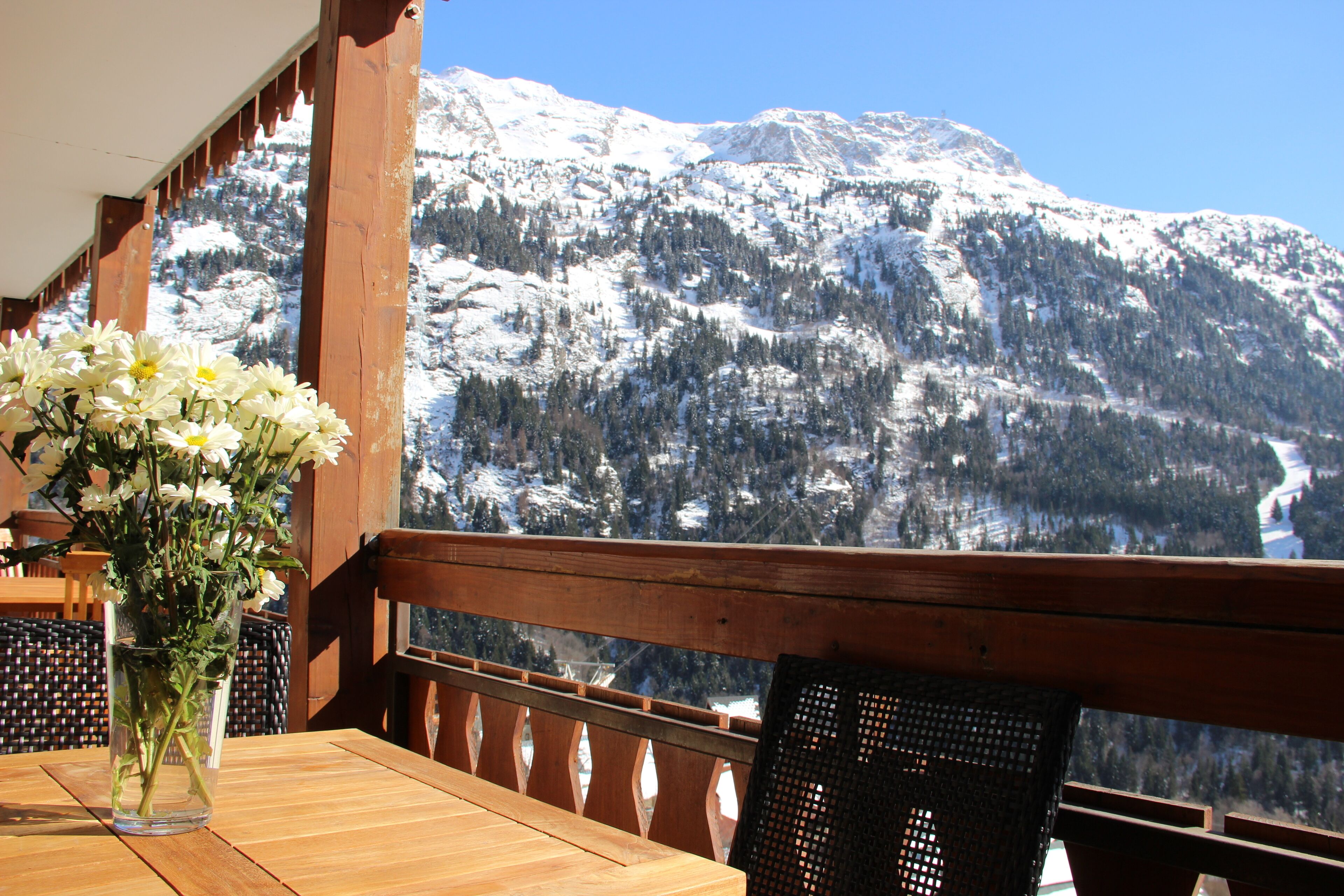 Ein Holztisch mit einer Vase voller weißer Blumen, ein Korbstuhl und ein Blick auf schneebedeckte Berge.