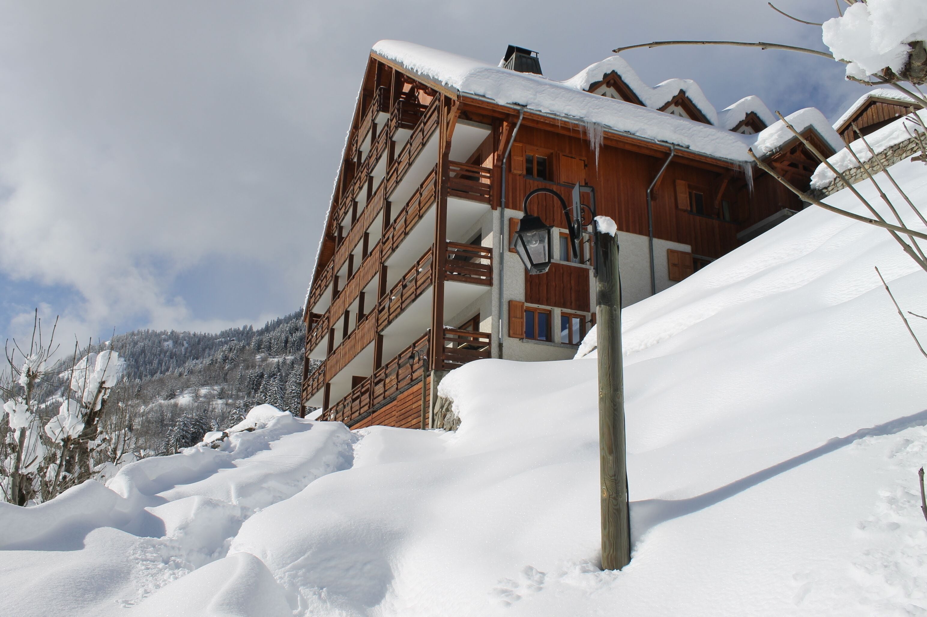 A wooden chalet with multiple balconies, surrounded by snow-covered trees and a mountainous backdrop.