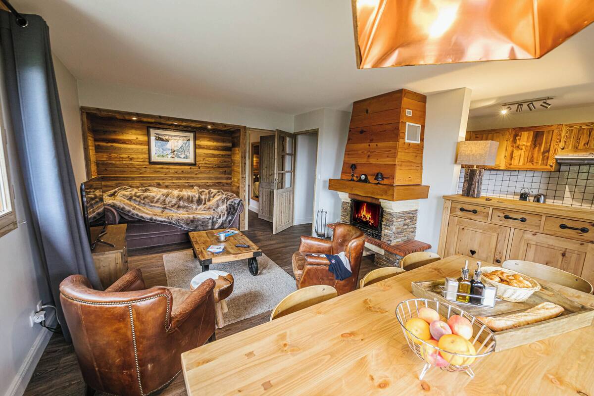 A cozy living room with a sofa, leather chairs, a wooden dining table with fruit, and a fireplace.
