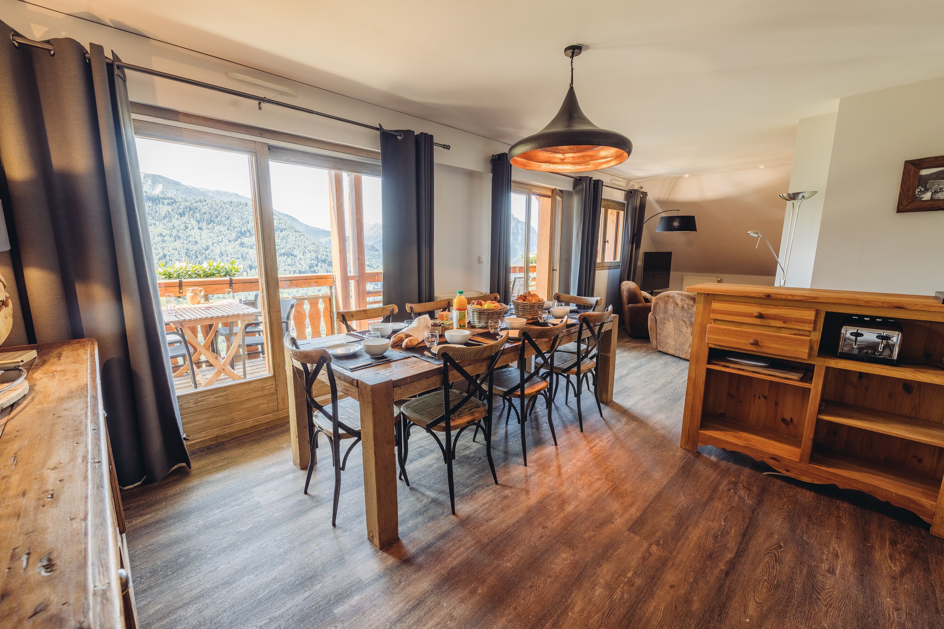 Zona pranzo con tavolo e sedie in legno, un lampadario e vista sulle montagne attraverso ampie finestre.