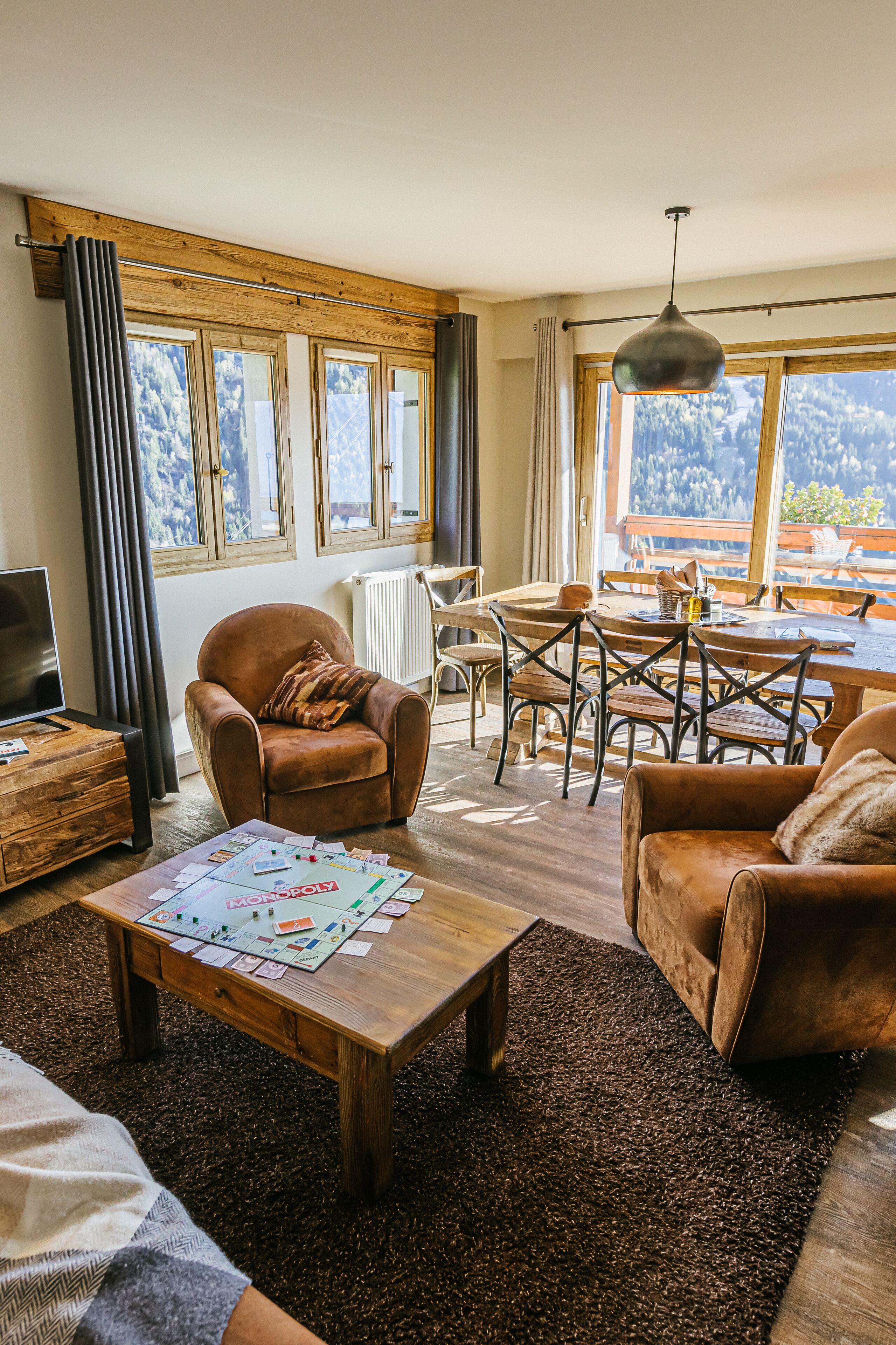 Un salon chaleureux avec un fauteuil en cuir marron, une table basse en bois sur laquelle sont posés des jeux de société, un coin repas avec une table et des chaises, et de grandes fenêtres offrant une vue sur l’extérieur.