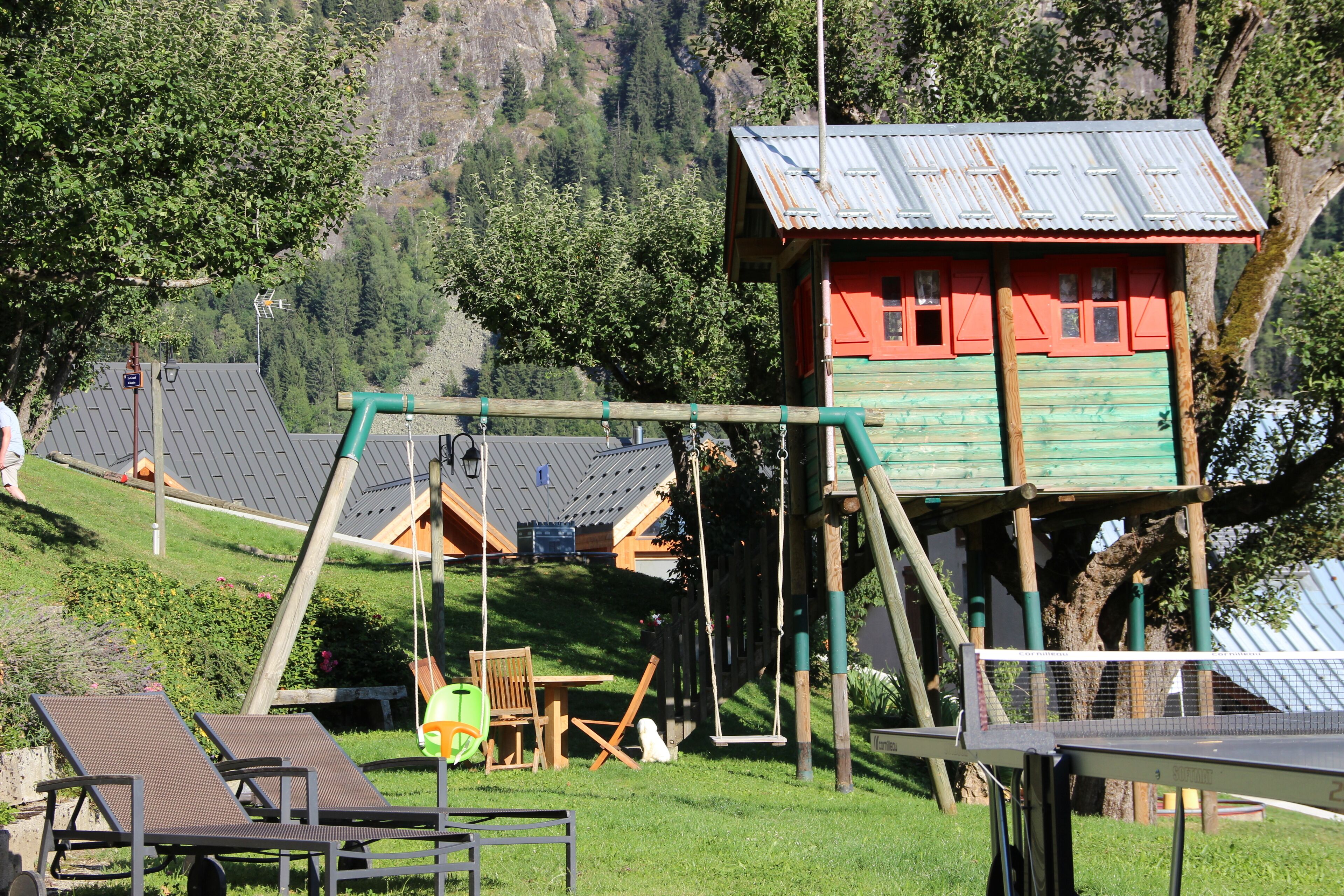 A wooden playhouse with a swing set, a small table, and a chair in a grassy area with a mountain backdrop.