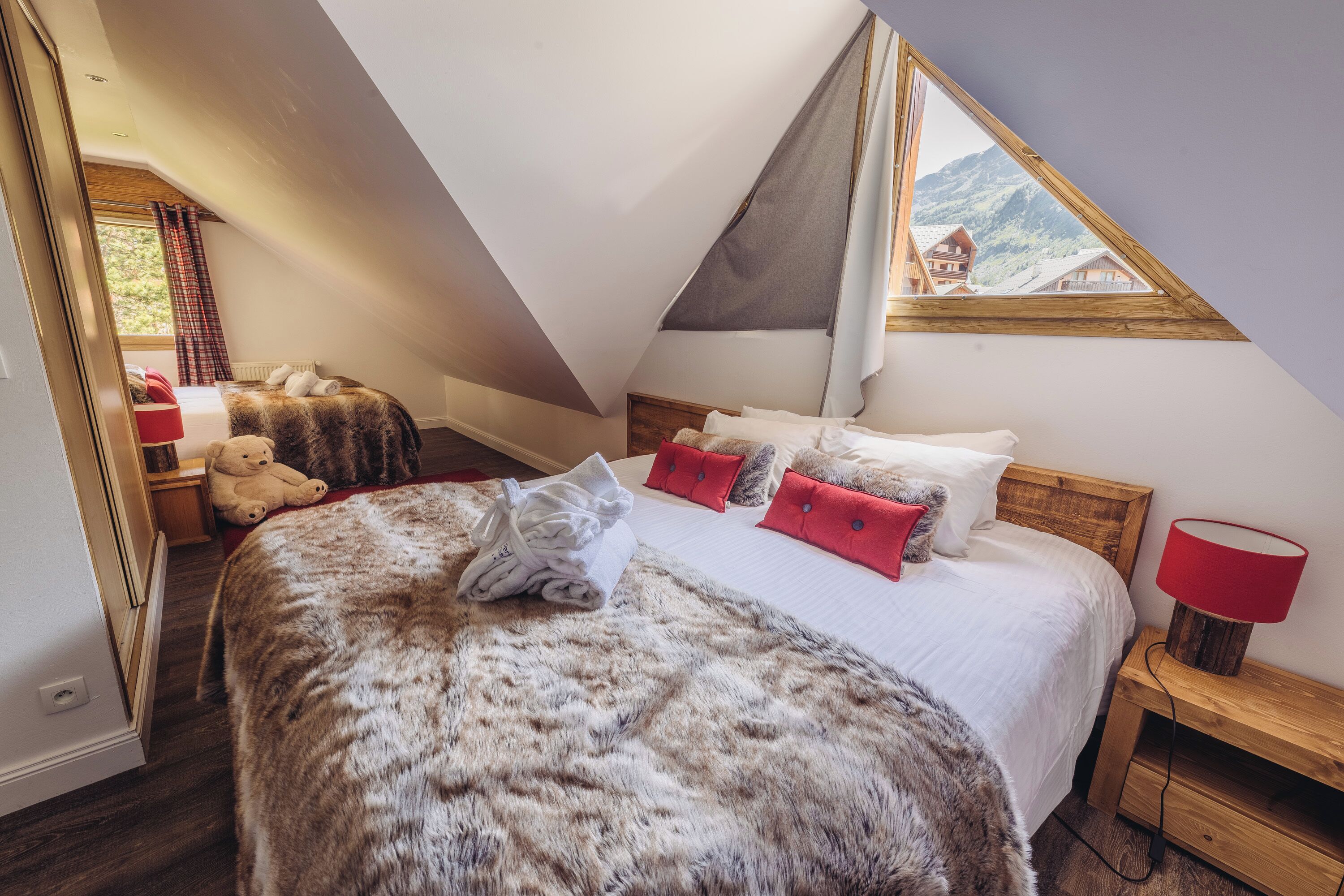 Un dormitorio con techo inclinado, una cama con una manta de piel, almohadas rojas y una mesita de noche con una lámpara.