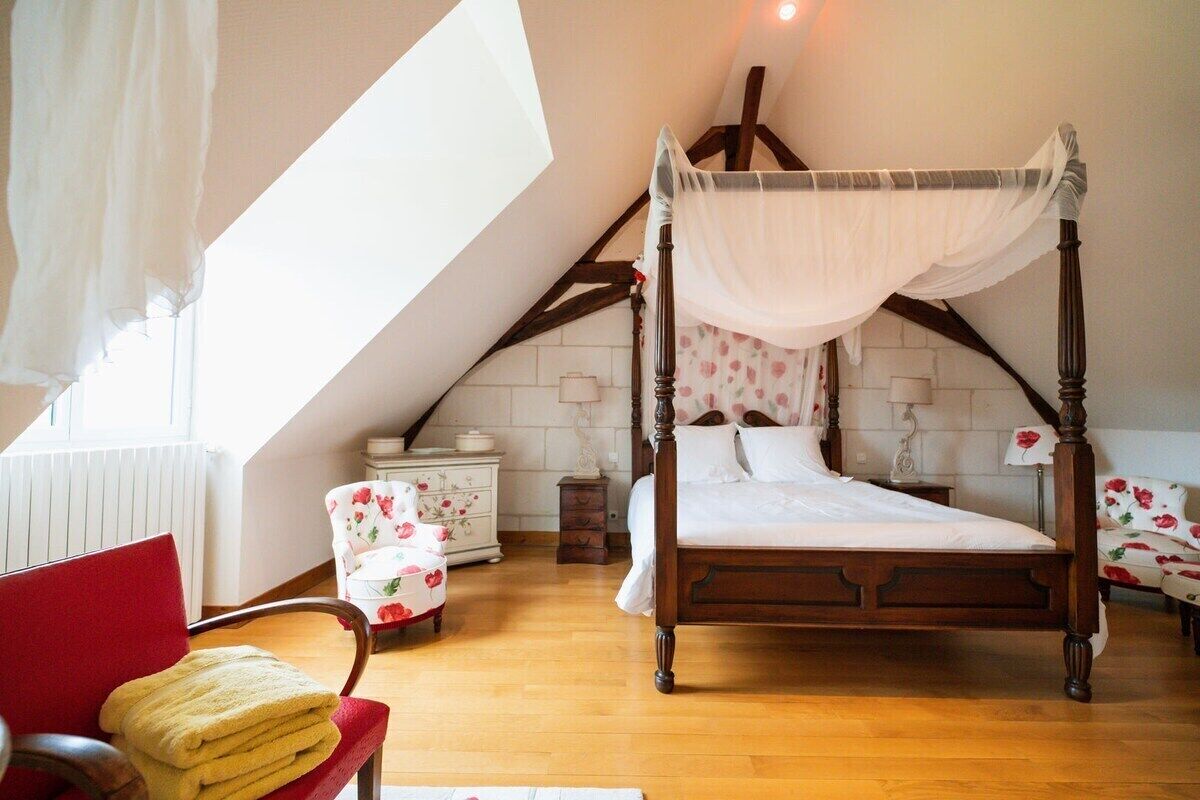 Une chambre à coucher avec un lit à baldaquin, un fauteuil rouge, une petite table de chevet et une fenêtre avec des rideaux.