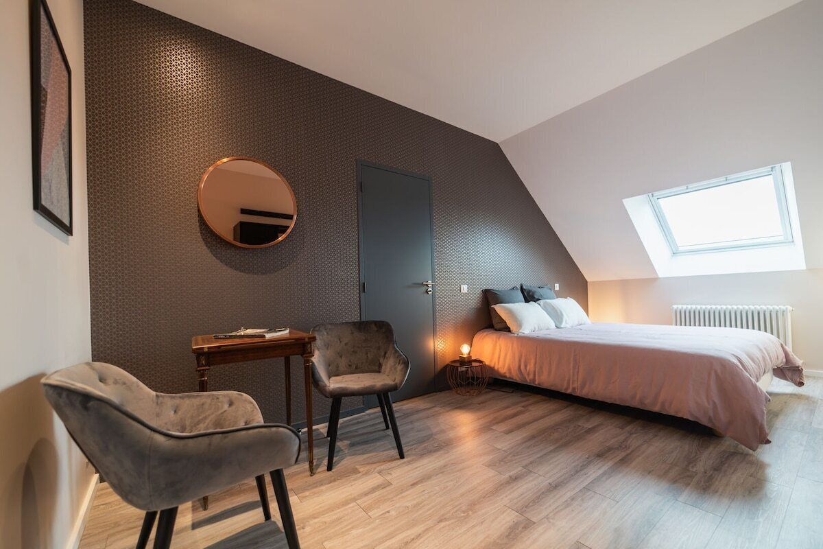 A modern bedroom with a sloped ceiling, a round mirror, a bed with a headboard, a desk, and two chairs.