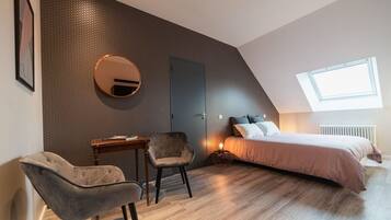 A modern bedroom with a sloped ceiling, a round mirror, a bed with a headboard, a desk, and two chairs.