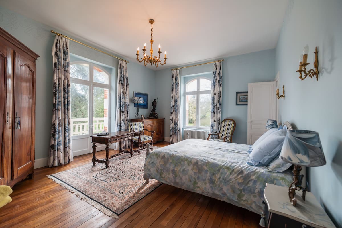 A bedroom with a large bed, wooden furniture, a chandelier, and a view of the outdoors.