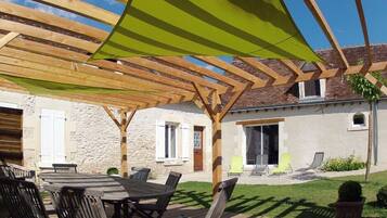 Area patio esterna con una pergola in legno, un tavolo rotondo con sedie e un prato con qualche sedia a sdraio.