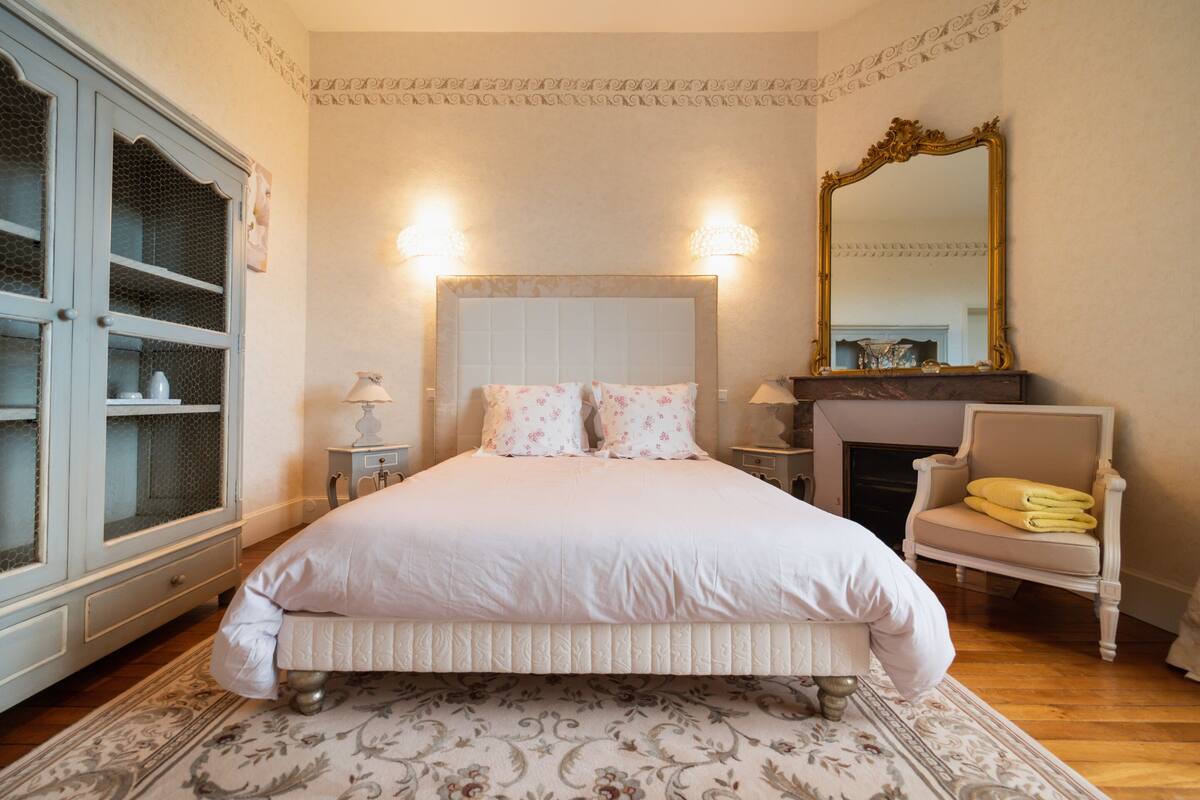 A bedroom with a large bed, a dresser, a chair, a mirror, and a fireplace.