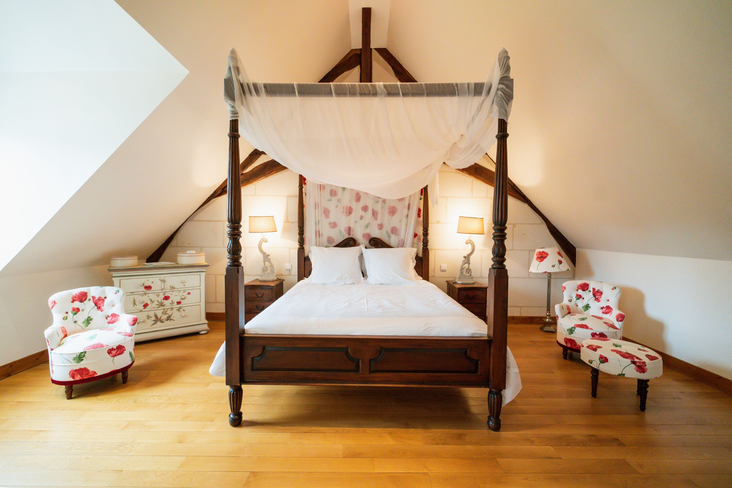 Una cama con dosel sobre cuatro postes, con estructura de madera y ropa de cama blanca, flanqueada por dos mesitas de noche a juego y dos sillones con estampado floral.