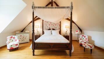 Una cama con dosel sobre cuatro postes, con estructura de madera y ropa de cama blanca, flanqueada por dos mesitas de noche a juego y dos sillones con estampado floral.