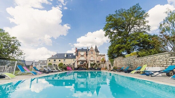 A swimming pool with lounge chairs and a stone wall, surrounded by trees and a large building with a pointed roof. - Domaine de la Poignardiere (Chatillon-sur-Indre)