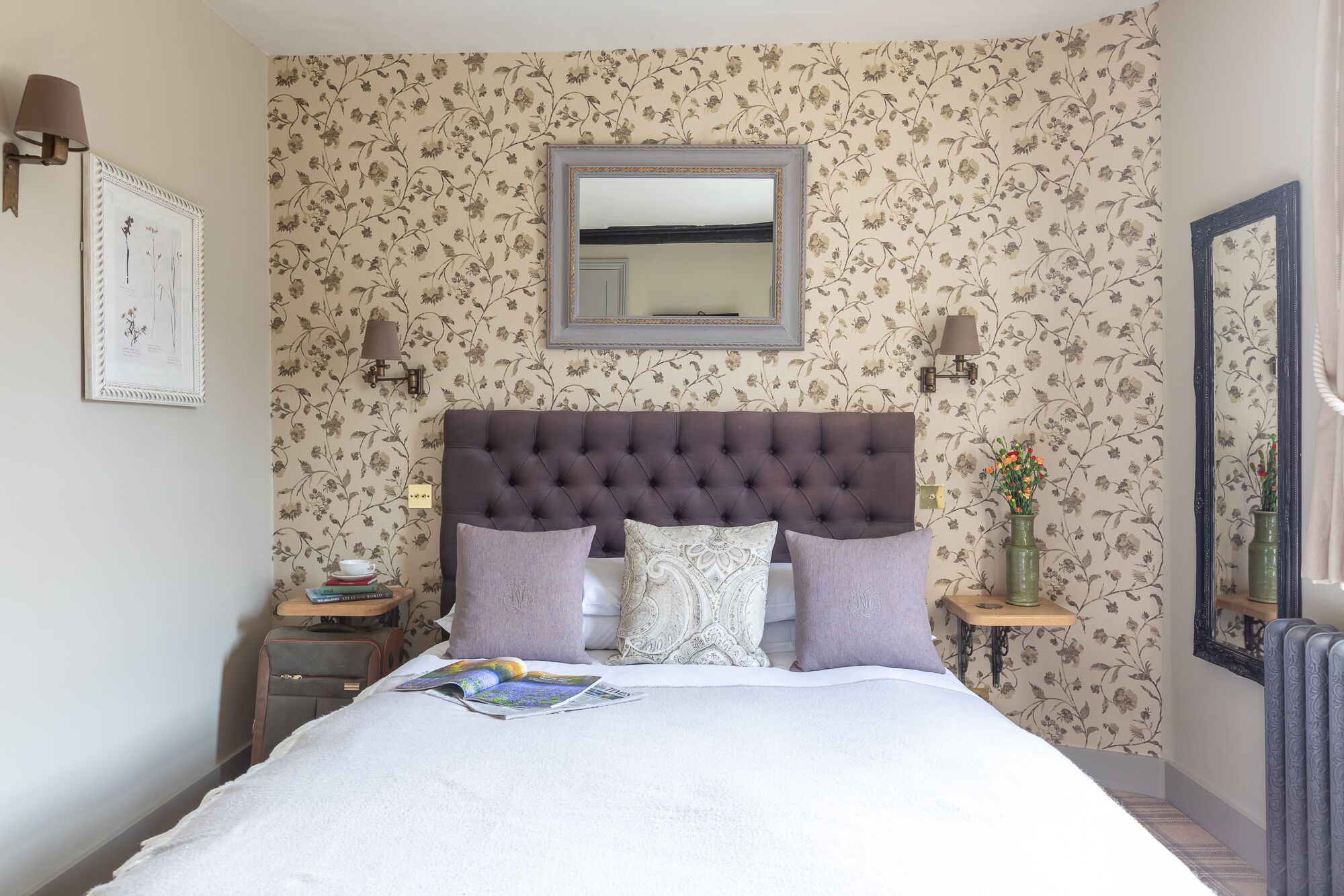 A neatly arranged bedroom with a tufted headboard, a mirror above the bed, floral wallpaper, and a bedside table with a lamp and a vase of flowers.