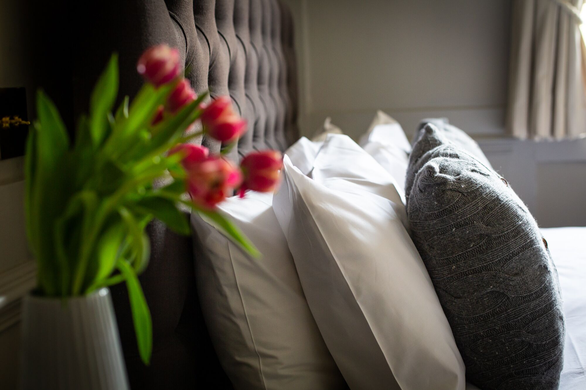 Una cama con almohadas blancas y grises, un cabecero oscuro y un jarrón con flores rojas.
