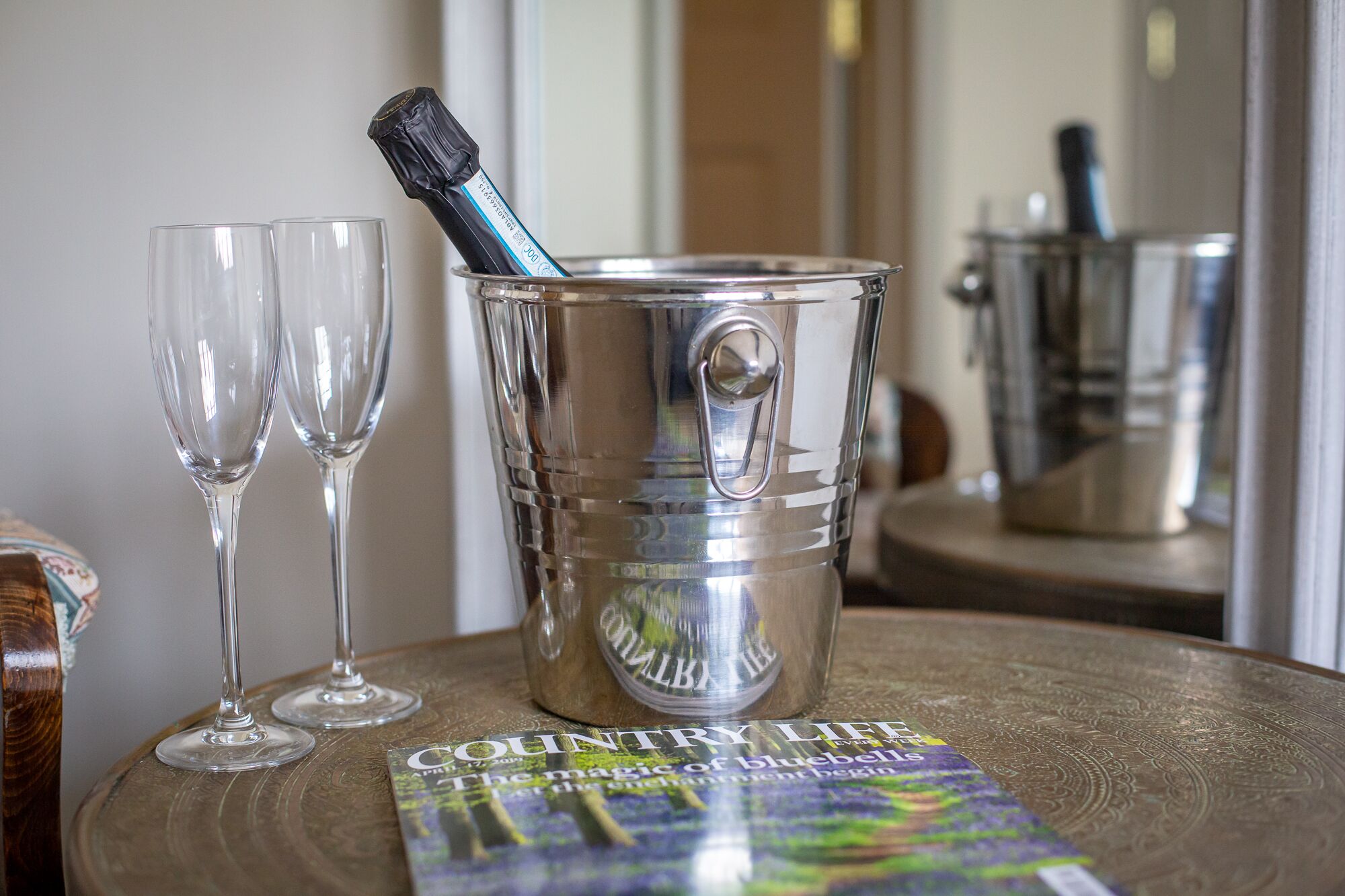 A table with two empty wine glasses, a metal bucket with a bottle of champagne, and a magazine titled "COUNTRY LIFE".