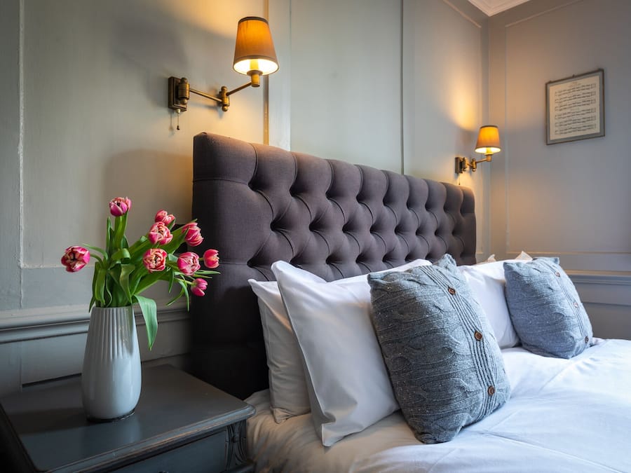 A neatly made bed with a tufted headboard, a vase of pink tulips, and two wall-mounted lamps.
