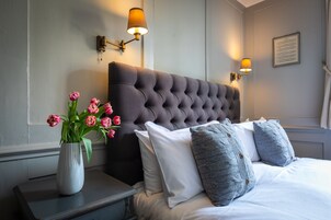 Una cama bien hecha con cabecera capitonada, un florero con tulipanes rosados y dos lámparas fijadas a la pared.