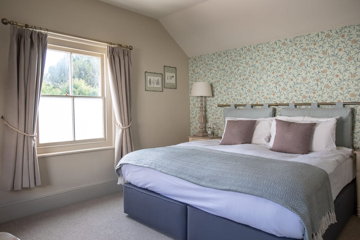 A bedroom with a large bed, a window with curtains, a nightstand with a lamp, and floral wallpaper.