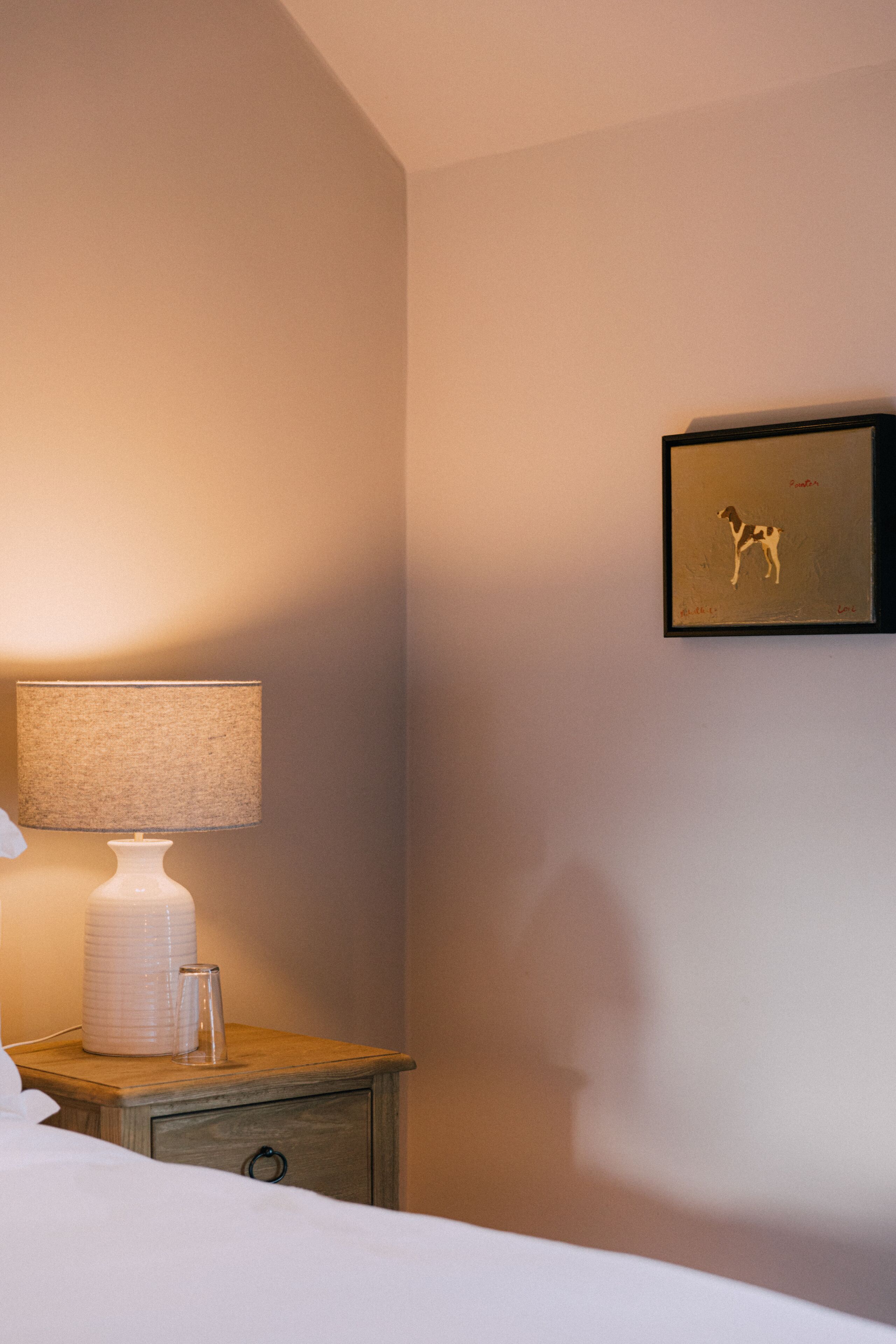 A neatly made bed with a white comforter, a bedside table with a lamp, and a framed picture on the wall.