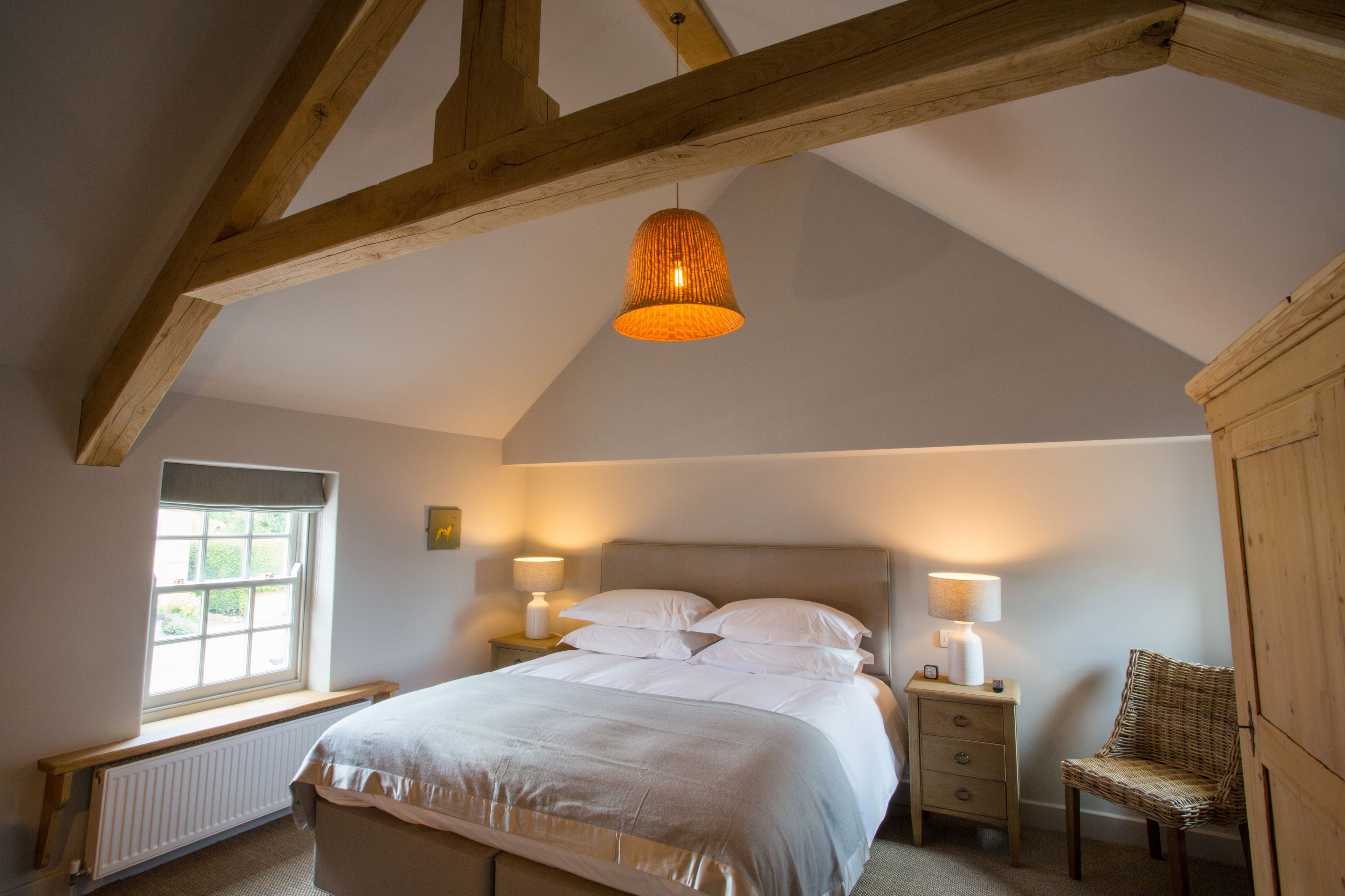 A bedroom with a large bed, two bedside tables, a chair, a window, and wooden beams on the ceiling.
