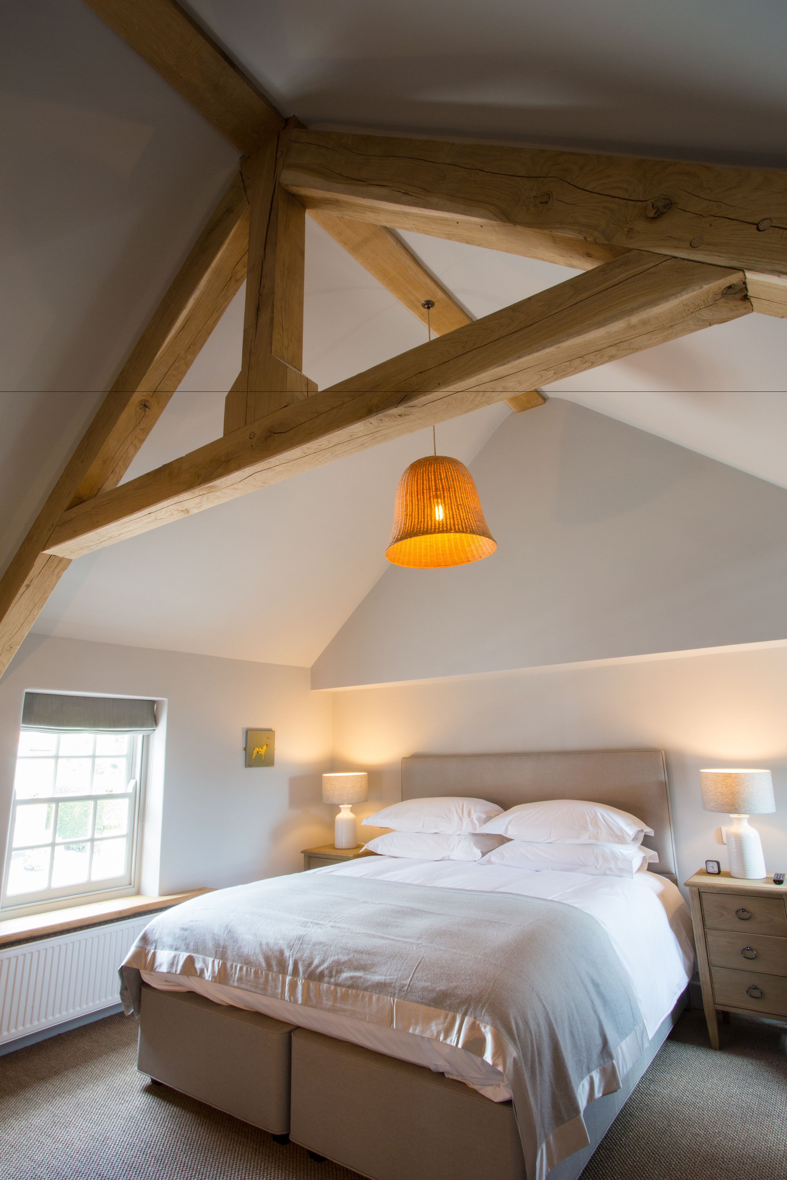 A bedroom with a wooden ceiling, a bed with white bedding, a bedside table with a lamp, and a window with a view of greenery.