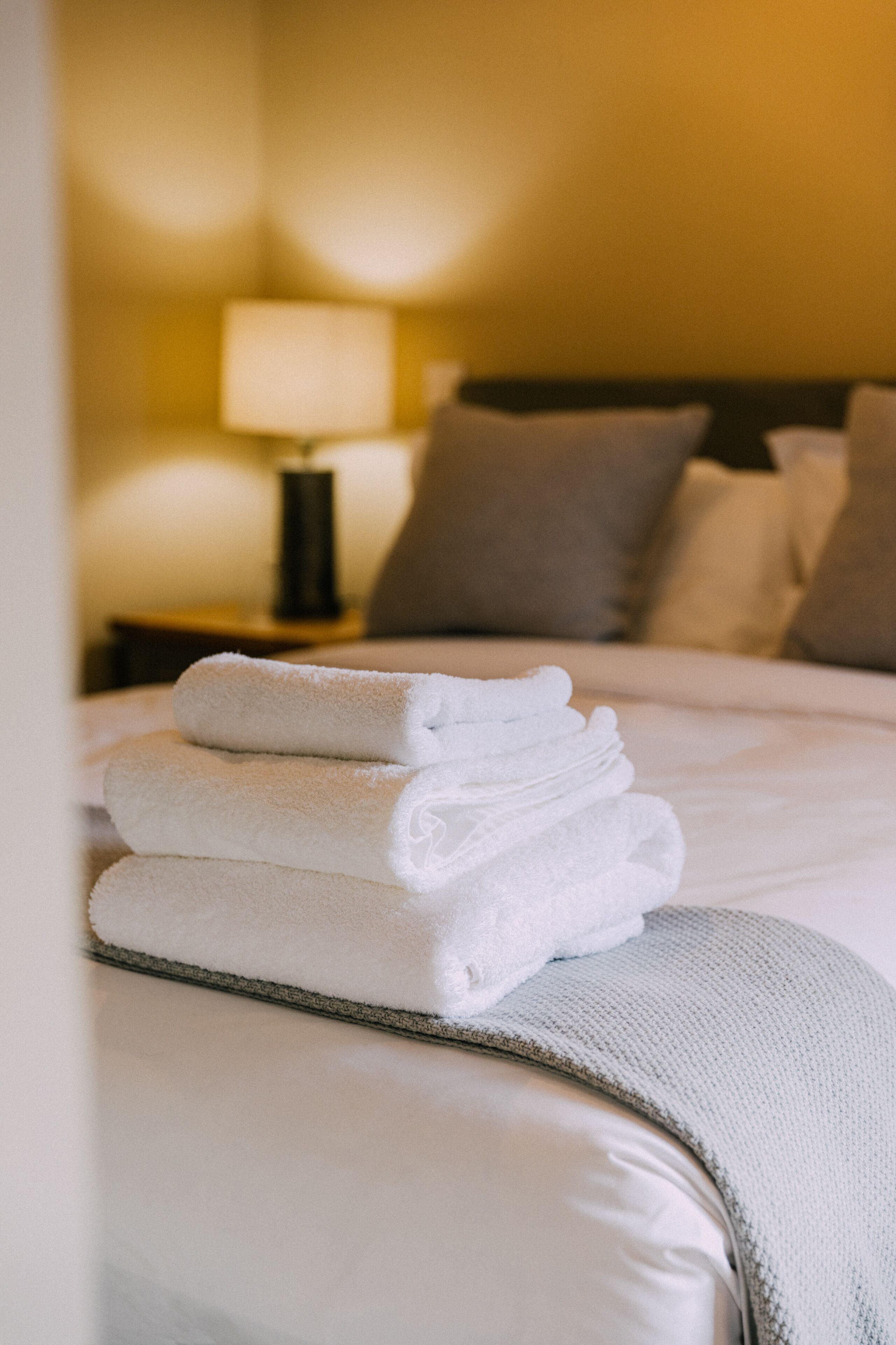 Een net opgemaakt bed met een stapel gevouwen witte handdoeken, een bedlamp en een nachtkastje met een lamp.