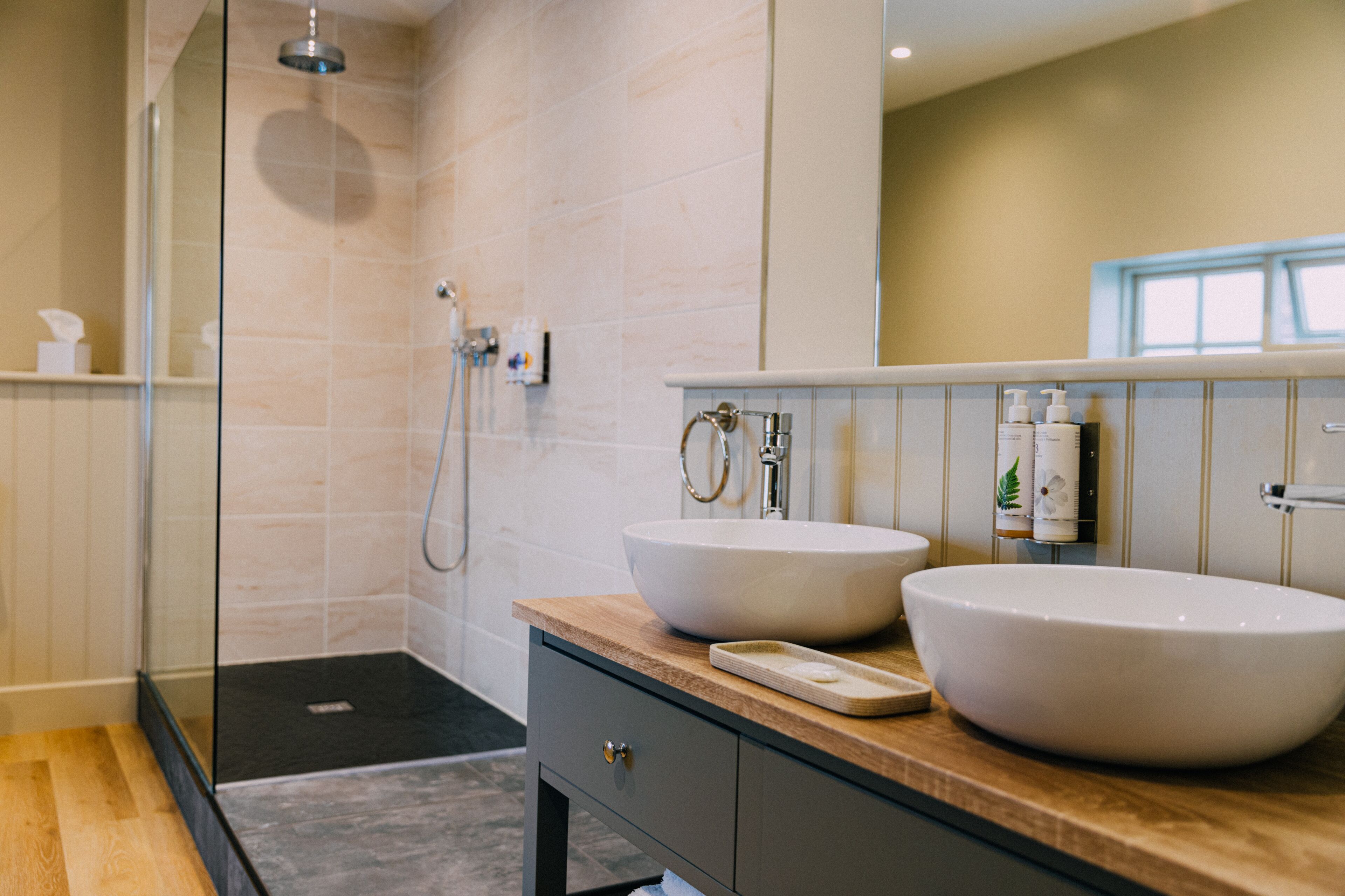 Un baño moderno con dos lavamanos grandes de color blanco, un box de ducha de cristal y un mueble de tocador de madera.