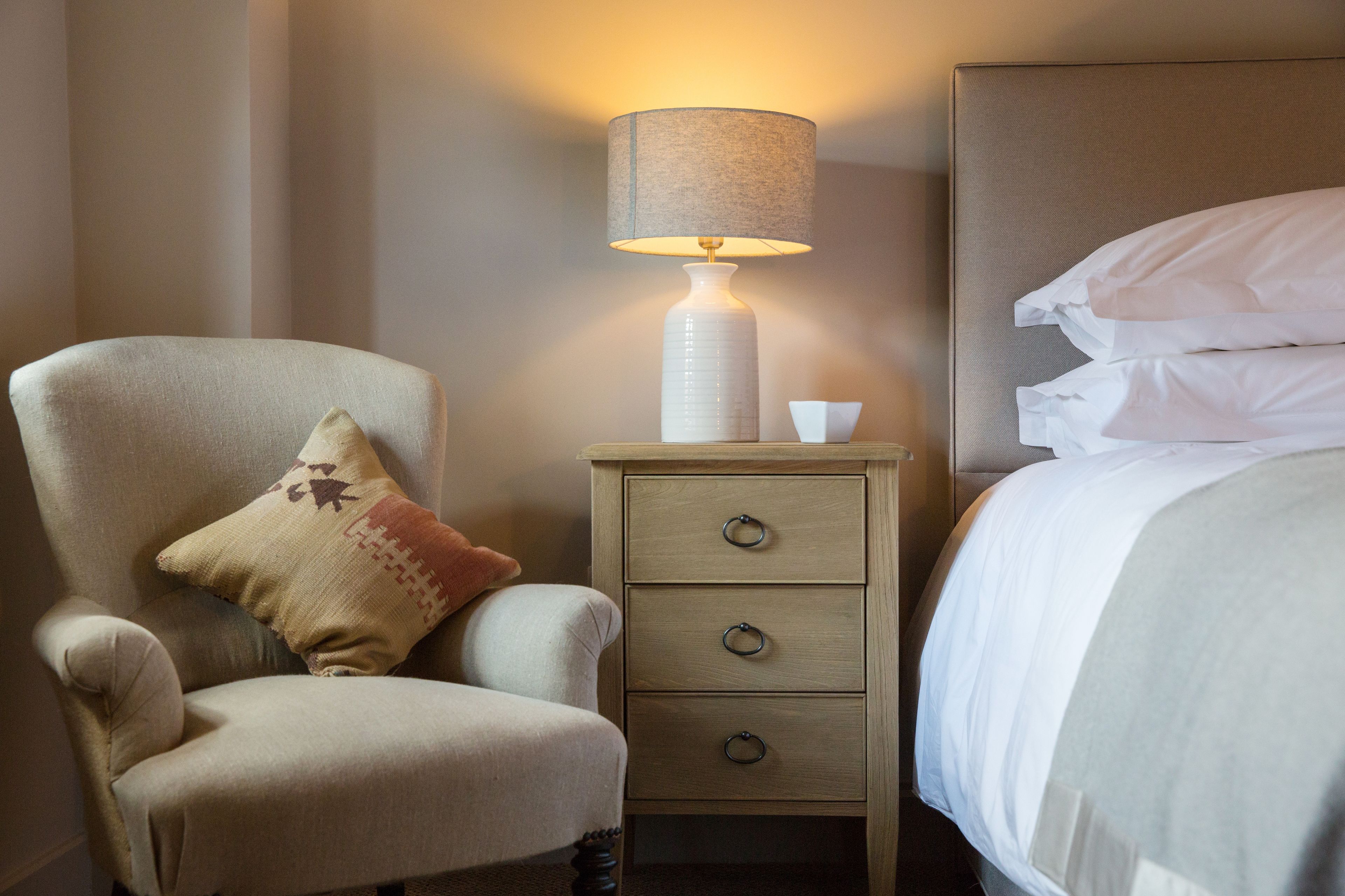 Una camera d'albergo con un letto, una poltrona con un cuscino, un comodino con una lampada e un mobiletto contenitore.