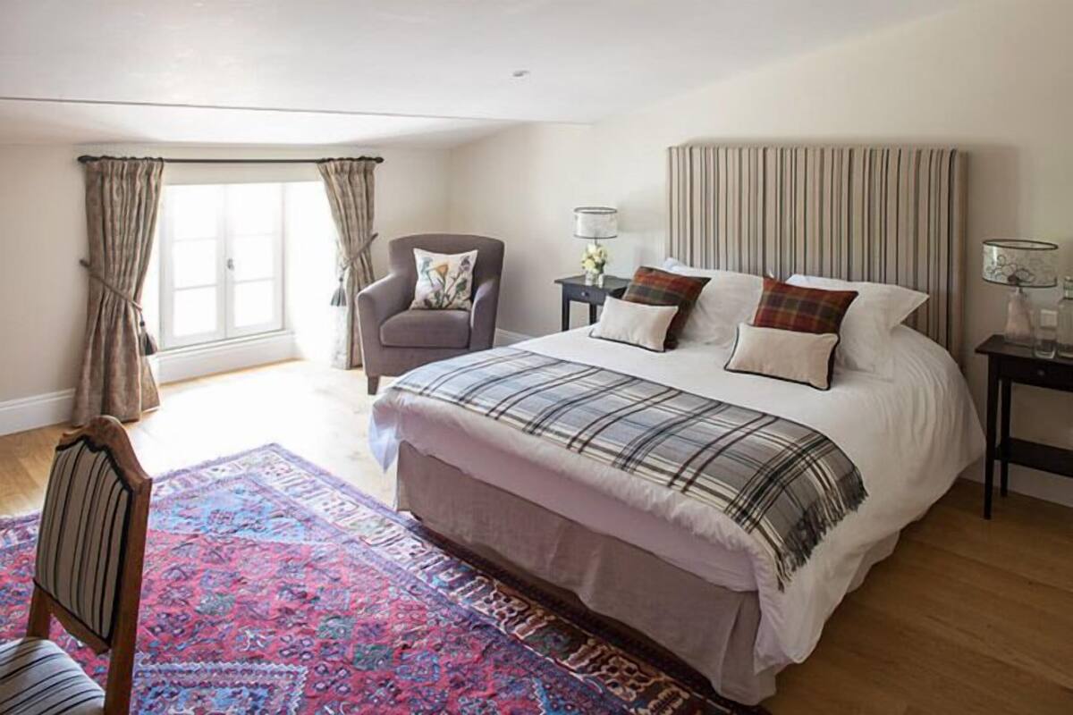 A bedroom with a large bed, a chair, a nightstand, a lamp, and a window with curtains.