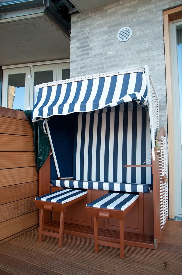 Una silla de playa con un toldo a rayas, una terraza de madera y un muro de ladrillo al fondo.