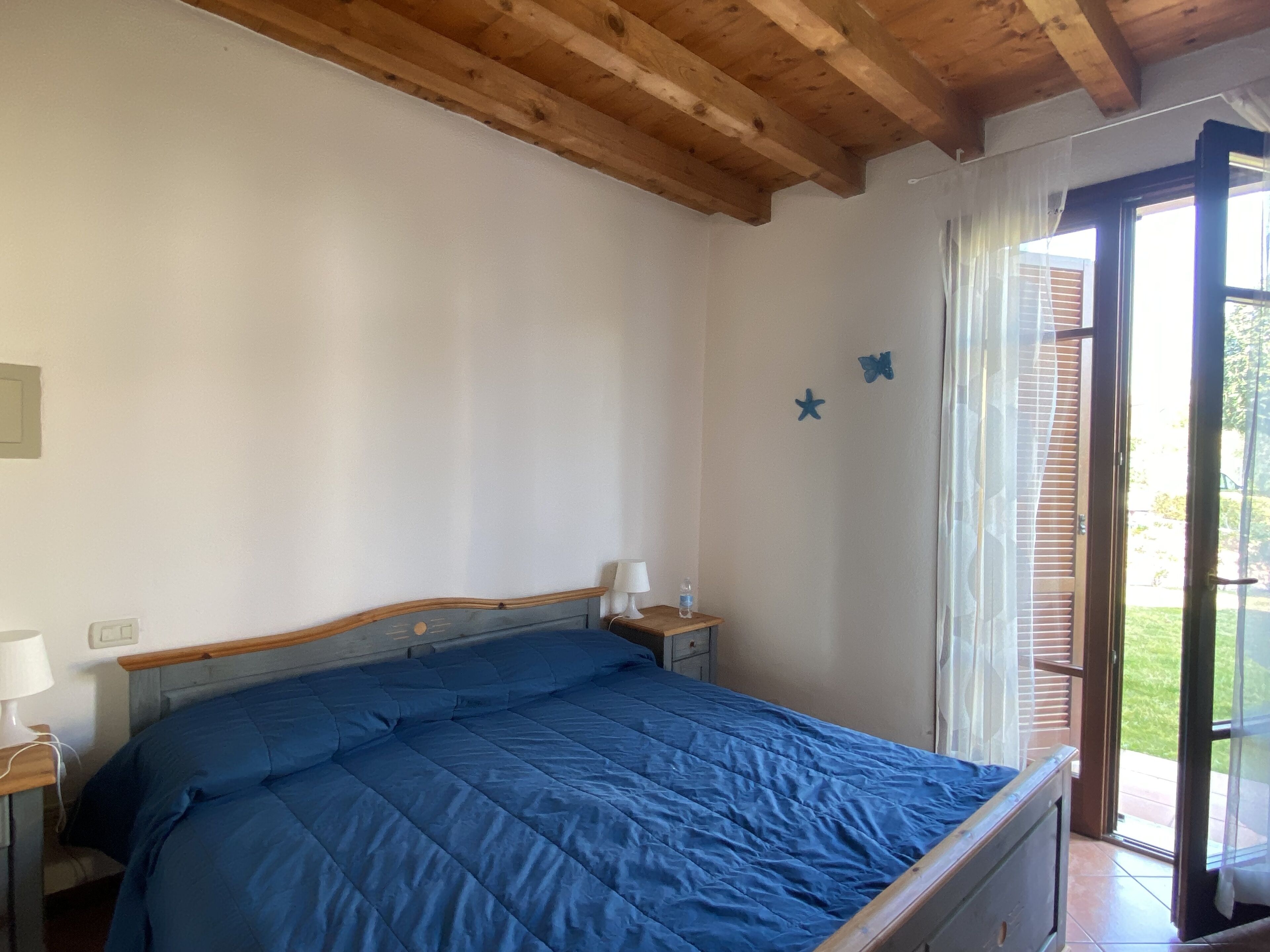A bedroom with a wooden ceiling, a bed with a blue bedspread, a nightstand with a lamp, and a window with blinds.