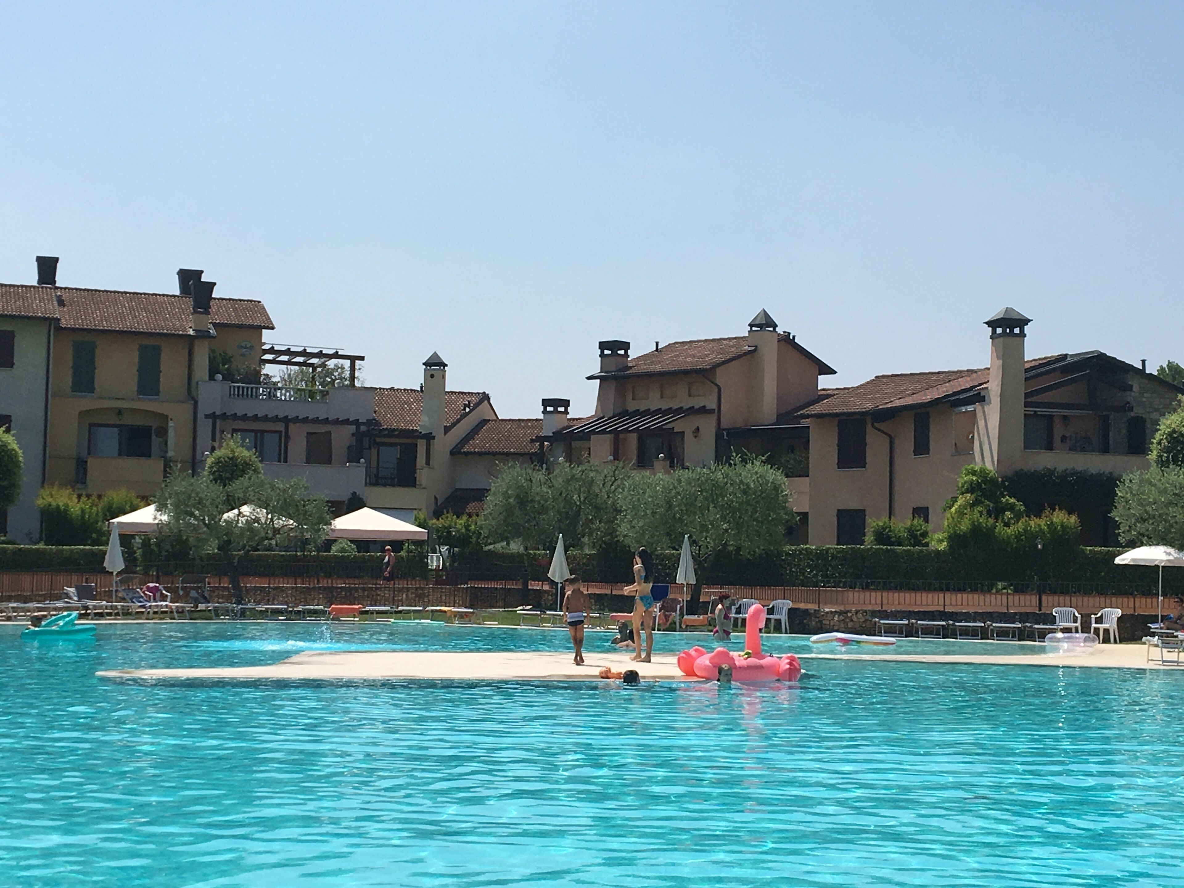 Una piscina con un dispositivo galleggiante rosa, circondata da una recinzione. Lungo il bordo piscina ci sono lettini e ombrelloni.