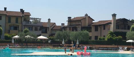Una piscina con un dispositivo galleggiante rosa, circondata da una recinzione. Lungo il bordo piscina ci sono lettini e ombrelloni.