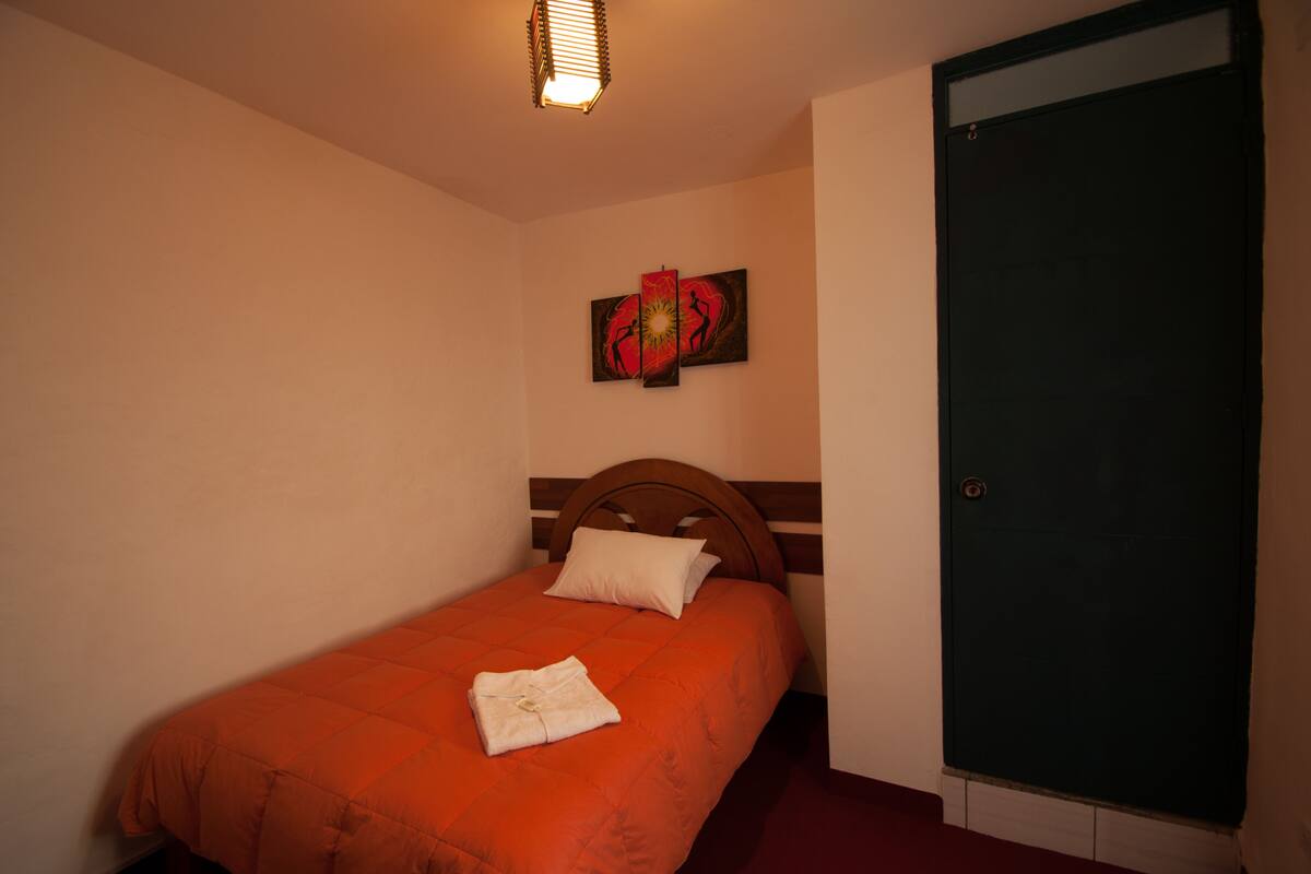 A single bed with an orange bedspread, a white pillow, and a folded white towel. There is a painting above the bed and a green door to the right.