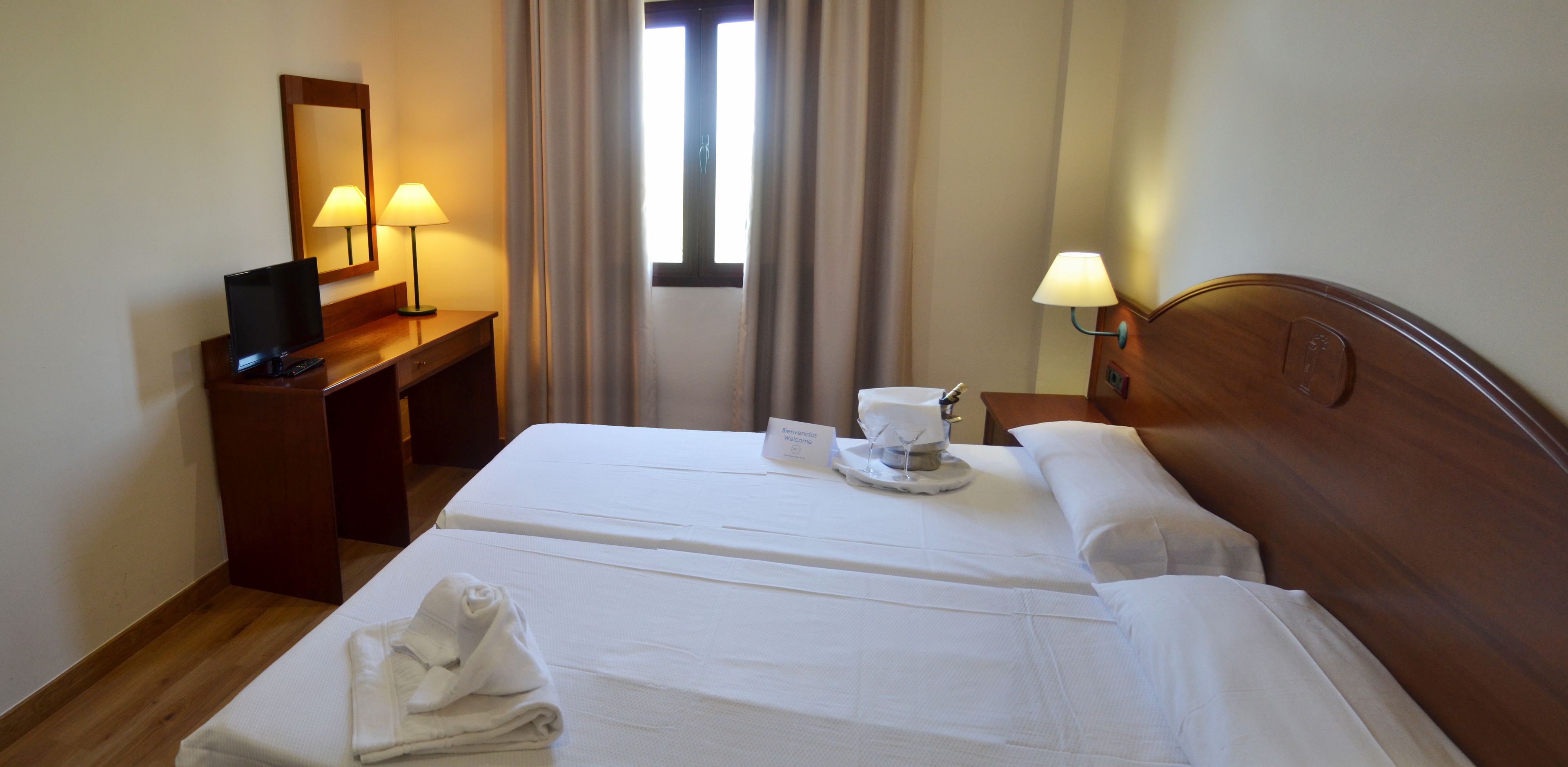 Habitació d'hotel amb un llit, un escriptori amb làmpada, un mirall i una finestra amb cortines.