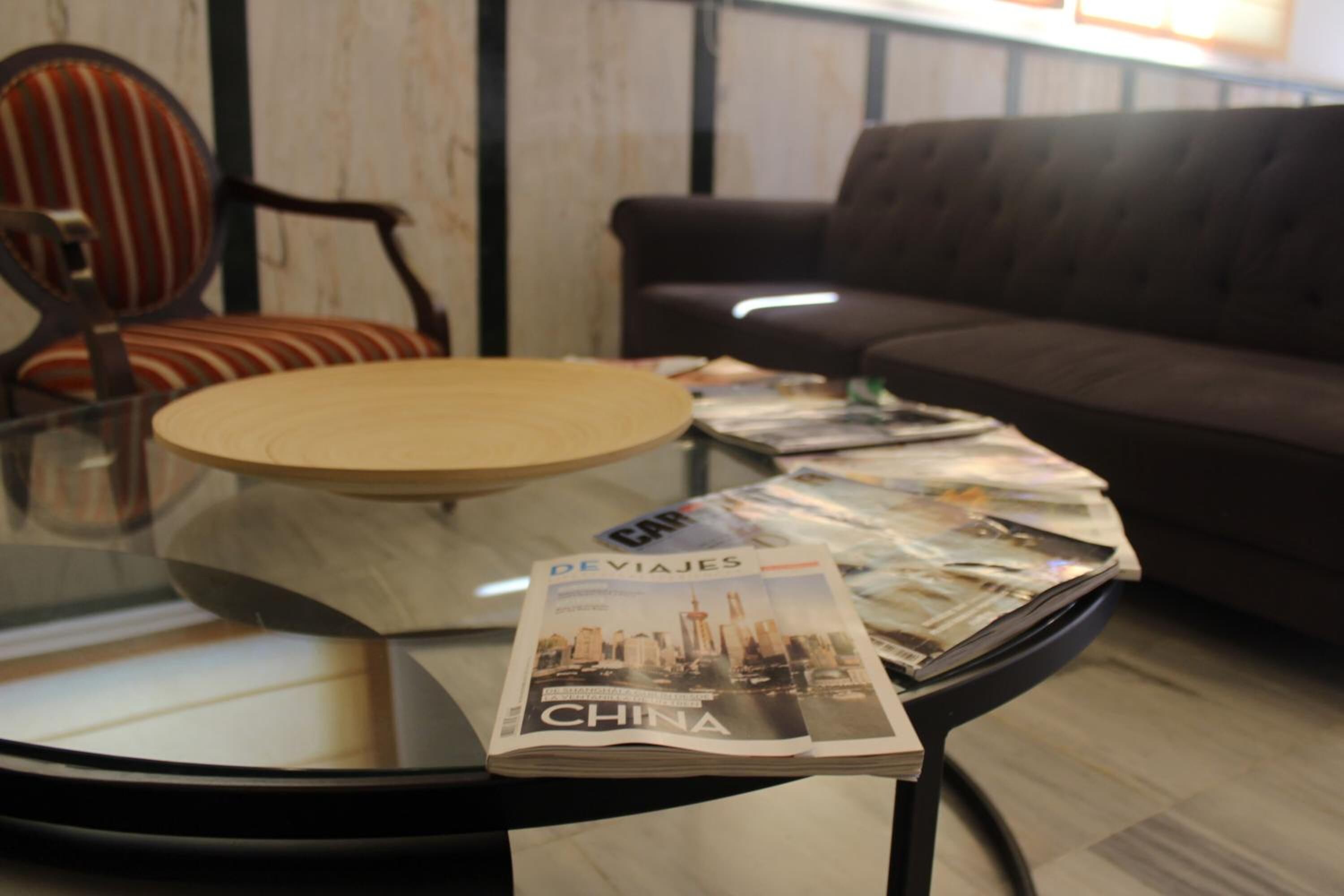 Taula de cafè amb tapa de vidre, amb una revista titulada "De Viajes" i "China", envoltada per un sofà marró i una cadira amb ratlles en una habitació amb panells de fusta.